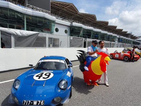 A man and woman stand beside a blue alpine antique car during the k-car global 2019 event by perodua, emphasizing the car's design and the occasion's atmosphere.