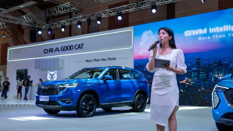 A woman stands next to a blue SUV during a GWM car launch event, emphasizing the vehicle's style and innovation.