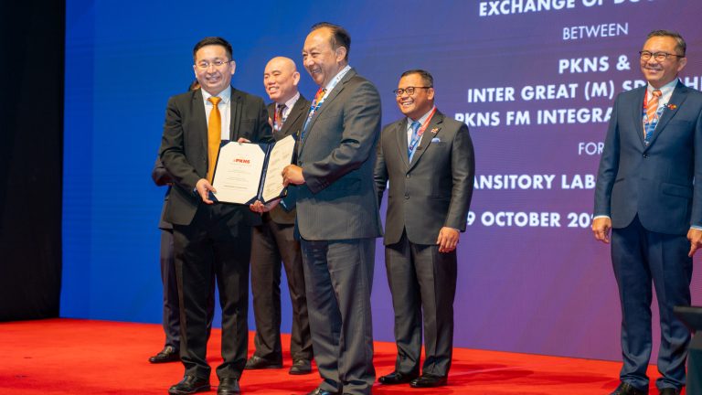 A group of men in suits gathered at an MoU event between PKNS and Inter Great (M) Sdn Bhd, where one man proudly presented the MoU certificate to another on a red carpeted stage with the Chief Minister of Selangor watching